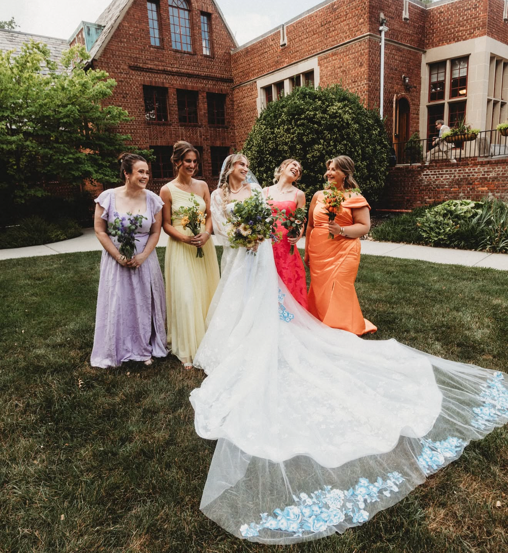 A social media post featuring a bride and five bridesmaids posing outdoors in front of a brick house. The bride wears a long white wedding gown with a long train, while the bridesmaids wear lavender, cream, and orange dresses. June bride with florals from Bits & Blooms Flower Farm.