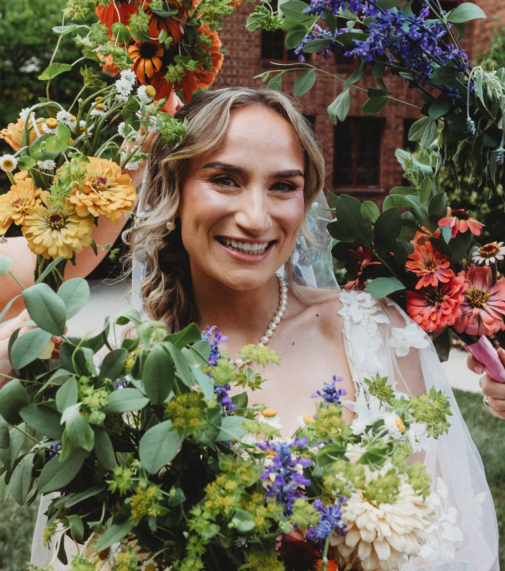 A social media story screenshot showing a smiling woman in a white bridal gown holding large, lush bouquets of green foliage, purple flowers, and orange blossoms in an outdoor garden setting. Bridal bouquet with wedding florals from Bits & Blooms Flower Farm.