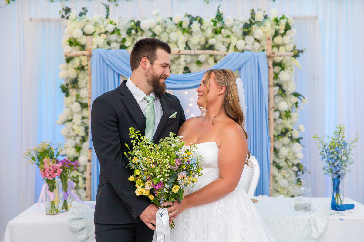 A bride in a white strapless wedding gown and a groom in a dark suit with a light green tie stand together in front of a decorated wedding backdrop featuring white flowers, greenery, and light blue drapery. The bride holds a colorful bouquet of wildflowers. Florals from Bits & Blooms Flower Farm.